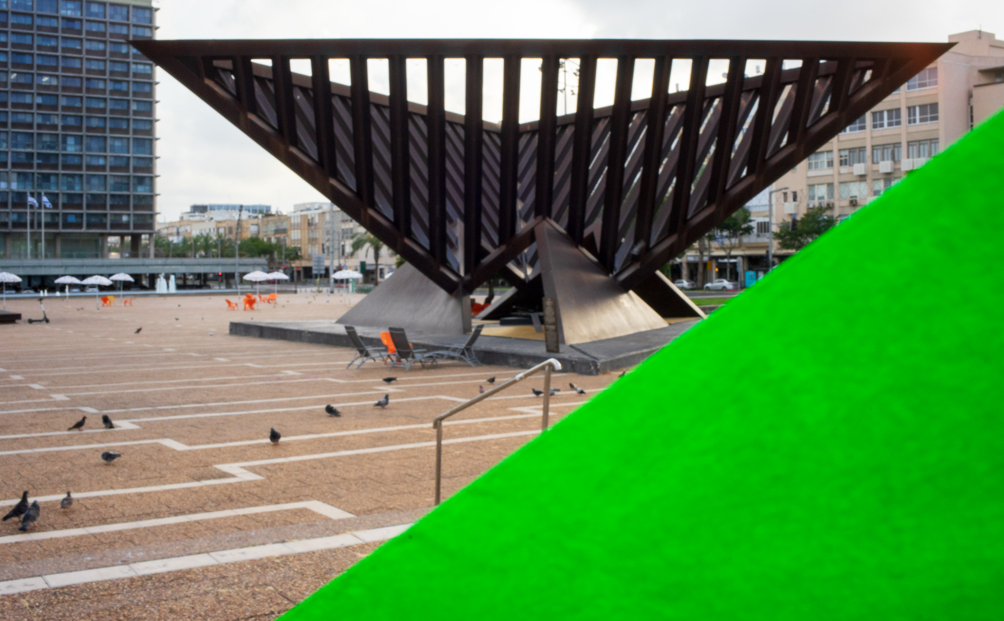 תמונה המציגה אנדרטה משולשת לשואה והתקומה מאת הפסל יגאל תומרקין, הממוקמת בכיכר רבין. חציו השמאלי-תחתון של התמונה מציג משולש בגוון ירוק זוהר המזוהה עם גווני הפרויקט.